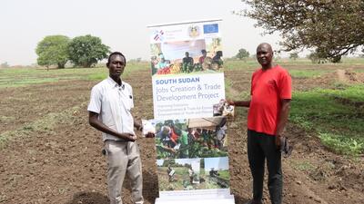 two south sudanese farmers stand next to banner in field